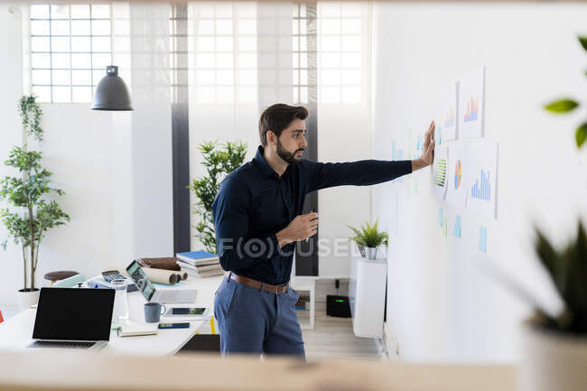 Joven hombre de negocios analizando estrategias mientras está parado contra la pared en la oficina - foto de stock