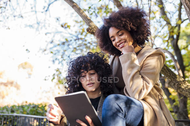 Молодая женщина показывает цифровой планшет улыбающейся подруге, сидящей с рукой на подбородке в парке — стоковое фото