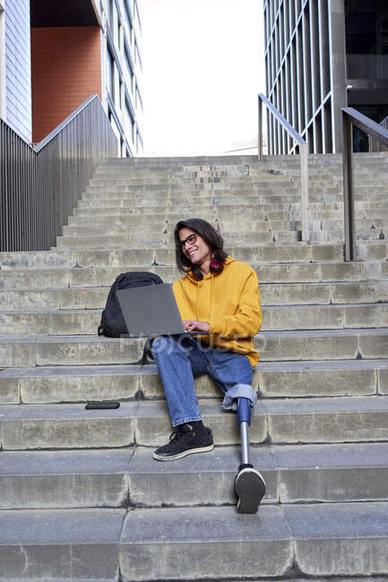 Усміхнений молодий каліка з штучною кінцівкою за допомогою ноутбука, сидячи на сходах у місті. — стокове фото
