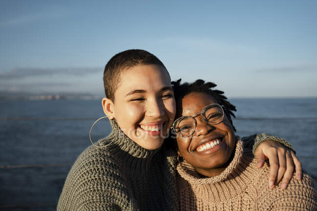 Amigas sonriendo sentadas contra el mar - foto de stock