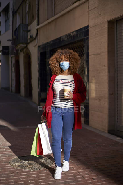 Young woman wearing protective face mask holding coffee cup and shopping bag while walking on footpath — Stock Photo