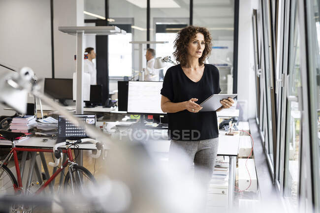 Empresaria sosteniendo tableta digital mientras está de pie con su colega en segundo plano en la oficina de planta abierta - foto de stock