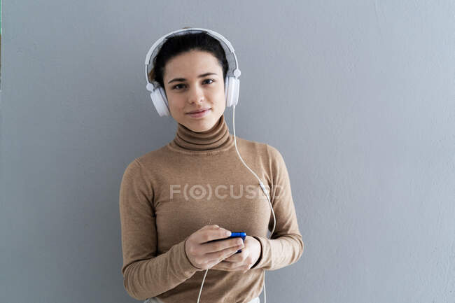 Giovane donna in possesso di smart phone durante l'ascolto di musica attraverso cuffie sullo sfondo grigio — Foto stock
