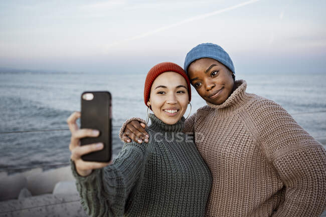 Amici che indossano cappello di maglia sorridente mentre prendono selfie attraverso il telefono cellulare mentre in piedi contro il mare — Foto stock