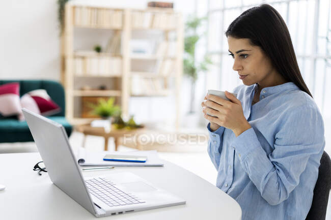 Femme entrepreneur boire du café tout en utilisant un ordinateur portable assis au bureau — Photo de stock