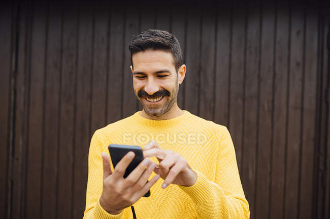 Sonriendo hombre adulto medio usando el teléfono móvil contra la pared de madera - foto de stock