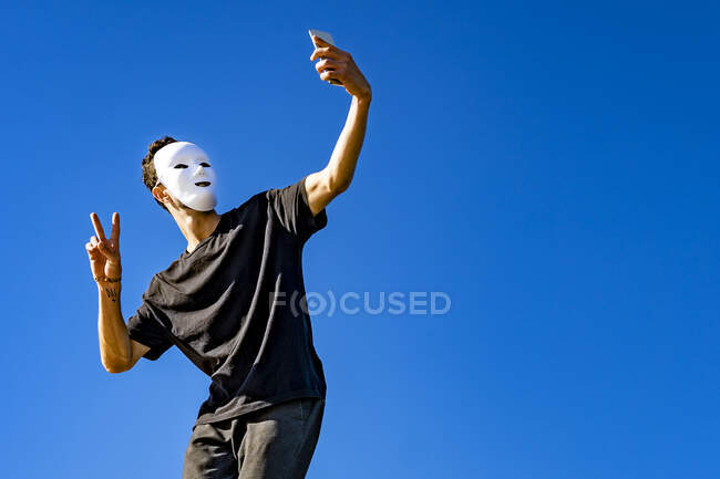 Hombre usando máscara blanca tomando selfie mientras que está parado contra cielo claro - foto de stock