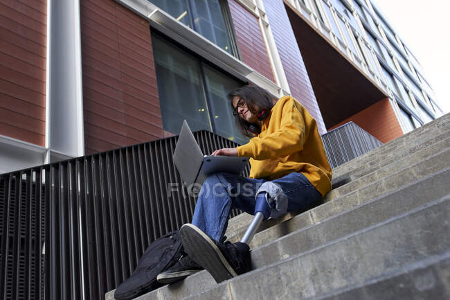 Jeune homme avec un membre artificiel à l'aide d'un ordinateur portable tout en étant assis sur des marches contre la construction — Photo de stock