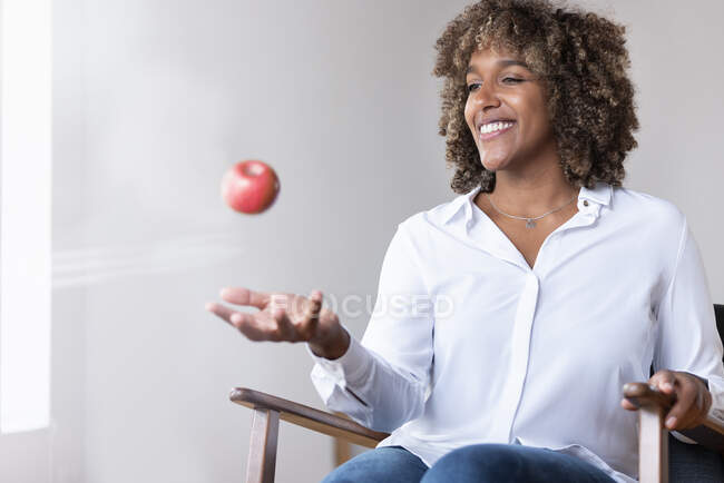 Femme souriante tout en jouant avec pomme assis sur un fauteuil à la maison — Photo de stock