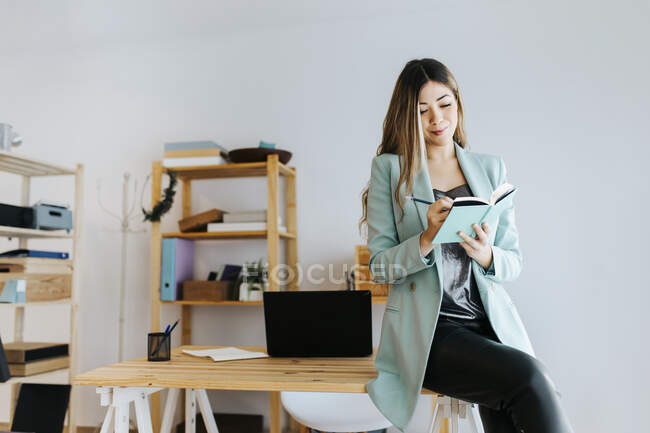 Donna d'affari sorridente che scrive nel diario mentre è seduta sulla scrivania contro il muro — Foto stock