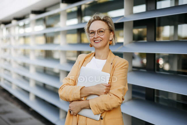 Sorridente maturo imprenditore femminile in possesso di laptop contro edificio per uffici — Foto stock
