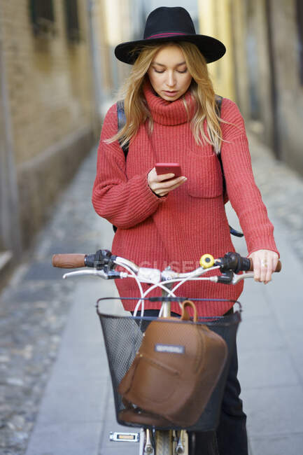 Young woman wearing hat using mobile phone while standing with bicycle on footpath — Stock Photo