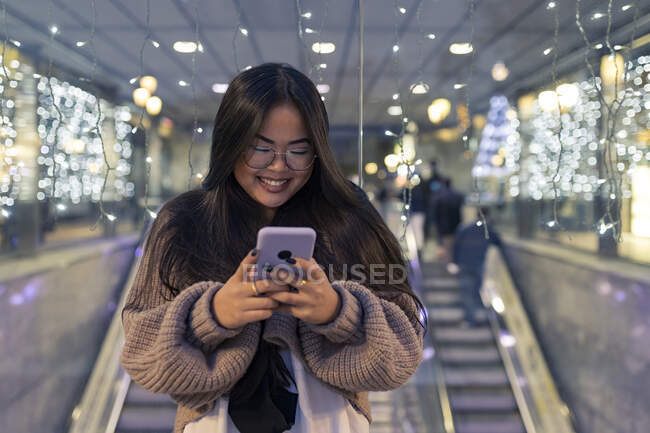 Mujer joven usando el teléfono inteligente mientras está de pie contra la pared de vidrio iluminado en el centro comercial - foto de stock