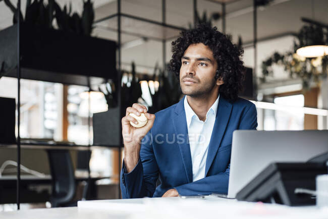 Pensativo joven empresario masculino mirando hacia otro lado mientras aprieta la bola de estrés en el escritorio en la oficina - foto de stock