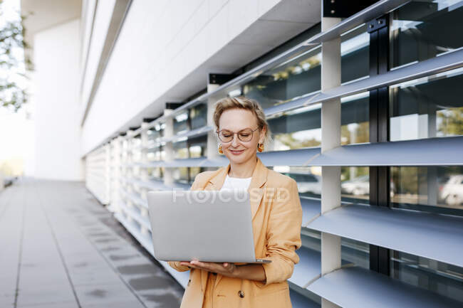 Emprendedora usando laptop mientras está de pie contra edificio de oficinas - foto de stock