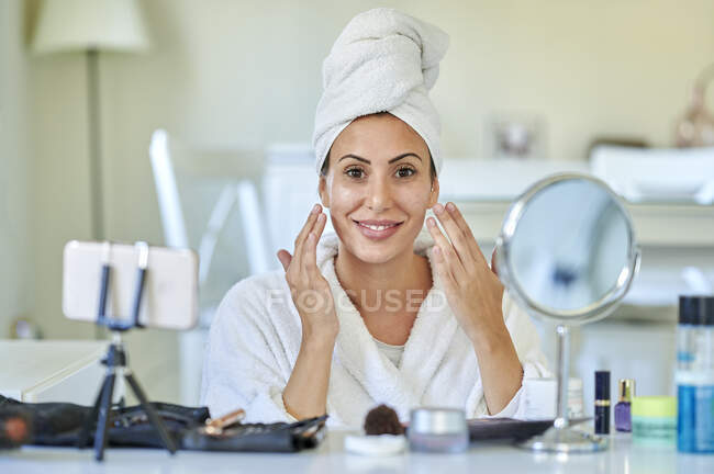 Blogueuse appliquant crème visage sur son visage tout en vlogging sur téléphone intelligent à la maison — Photo de stock