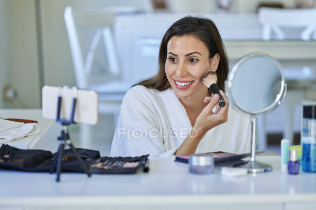 Улыбающаяся женщина-влиятель рассказывает о макияже на смартфоне дома — стоковое фото