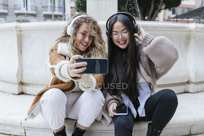 Sorridente amiche femminili prendere selfie indossando cuffie mentre seduto contro fontana — Foto stock