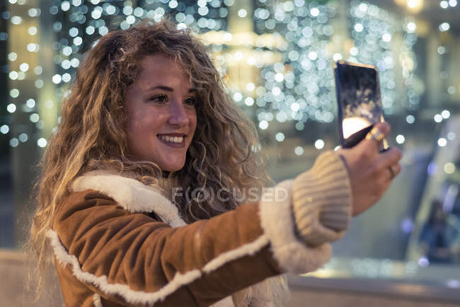 Молода жінка з холодним волоссям робить самовідтворення через смартфон проти освітленої скляної стіни — стокове фото