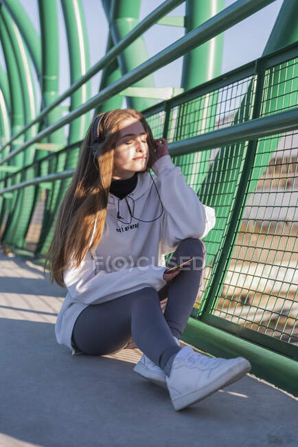 Девочка в наушниках слушает музыку, сидя на мосту — стоковое фото