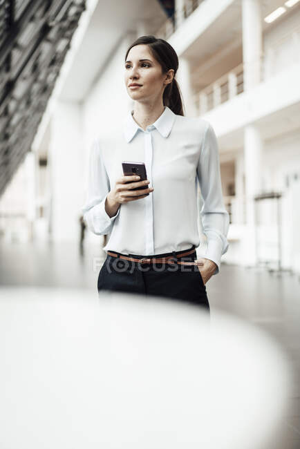 Confident businesswoman with smart phone looking away while standing in corridor — Stock Photo