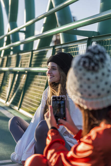 Mulher usando chapéu de malha tirando foto de amigo sorridente através do telefone celular enquanto sentado na ponte — Fotografia de Stock