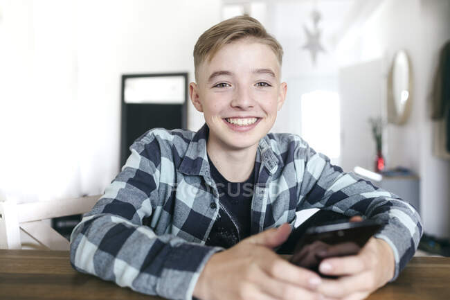 Счастливый мальчик, держа в руках мобильный телефон, сидя дома — стоковое фото