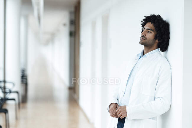 Meditado joven médico varón apoyado en la pared blanca en el pasillo del hospital - foto de stock
