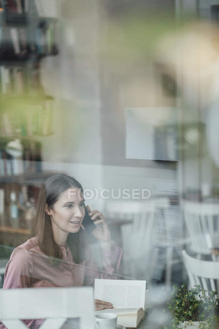 Businesswoman with book talking on smart phone in cafe — Stock Photo