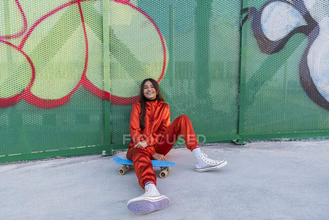 Молода жінка з скейтбордом посміхається, сидячи навпроти паркану графіті. — стокове фото