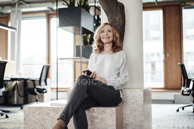 Smiling businesswoman with smart phone sitting on retaining wall in creative office — Stock Photo