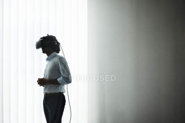 Joven emprendedor con auriculares de realidad virtual por cortina blanca en la oficina - foto de stock