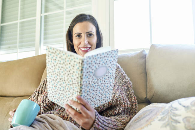 Donna sorridente con coppa che tiene libro sul divano a casa — Foto stock