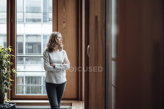 Femme entrepreneur avec les bras croisés regardant par la fenêtre dans le bureau — Photo de stock