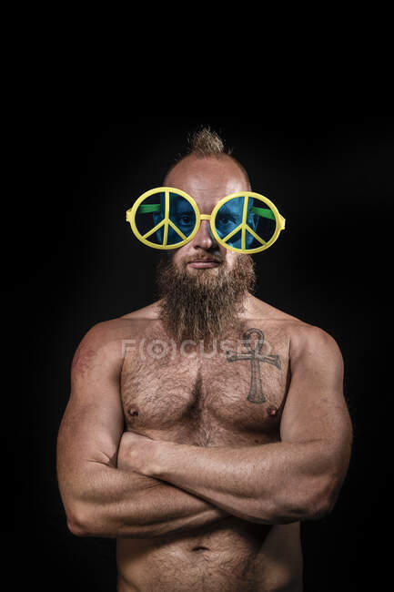 Shirtless male hipster wearing peace symbol novelty glasses standing with arms crossed against black background — Stock Photo
