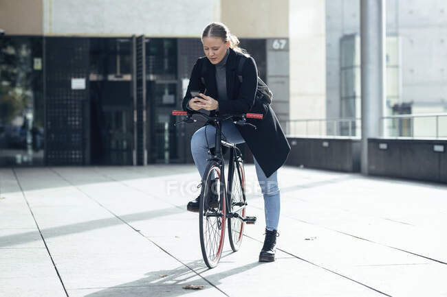 Junge Frau benutzt Handy auf Fahrrad vor Gebäude — Stockfoto