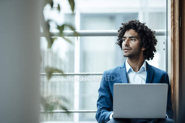 Pensativo profesional masculino mirando hacia otro lado mientras está sentado con el ordenador portátil contra la ventana en el lugar de trabajo - foto de stock