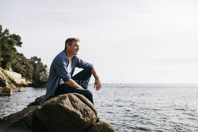 Nachdenklicher reifer Mann sitzt auf einem Felsen vor klarem Himmel — Stockfoto