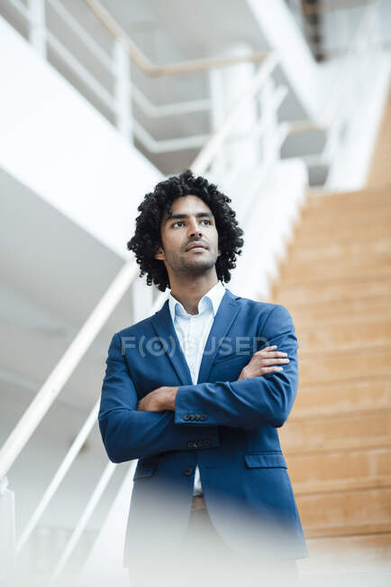 Homme d'affaires réfléchi avec les bras croisés contre l'escalier tout en regardant loin dans le bureau — Photo de stock