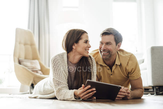 Smiling man and woman with digital tablet looking at each other while lying in home — Stock Photo