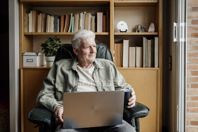 Uomo anziano guardando lontano mentre tiene il computer portatile a casa — Foto stock