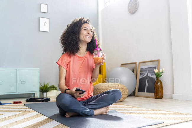 Весела жінка сидить з мобільним телефоном на килимку у вітальні — стокове фото