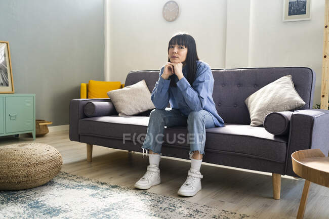 Молода жінка з рукою на підборідді сидить на дивані у вітальні вдома — стокове фото