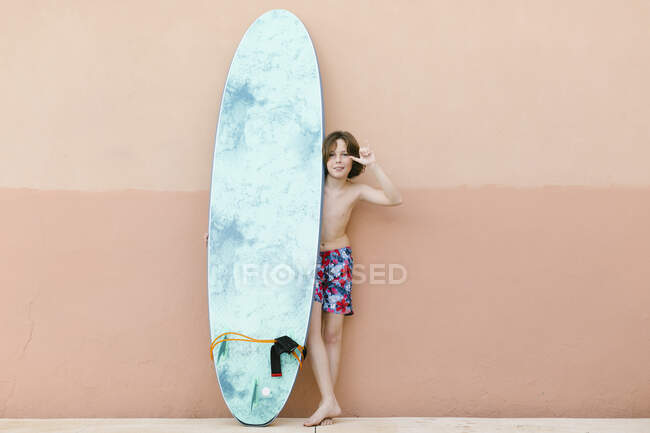 Ragazzo che indossa costume da bagno gesticolando mentre in piedi con tavola da surf davanti alla parete beige — Foto stock