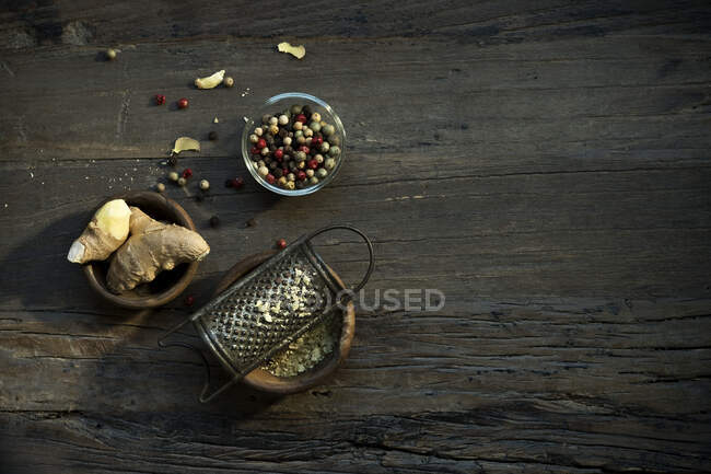 Tigelas com raiz de gengibre, ralador velho e vários grãos de pimenta deitados na superfície de madeira — Fotografia de Stock