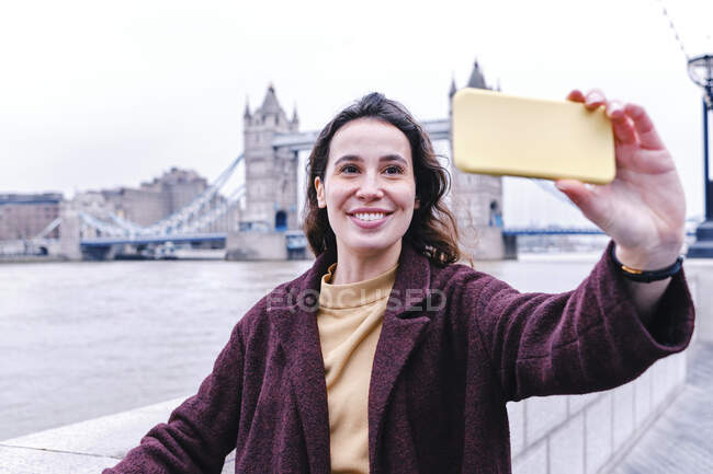 Sorridente giovane donna che prende selfie con Tower Bridge attraverso lo smart phone — Foto stock