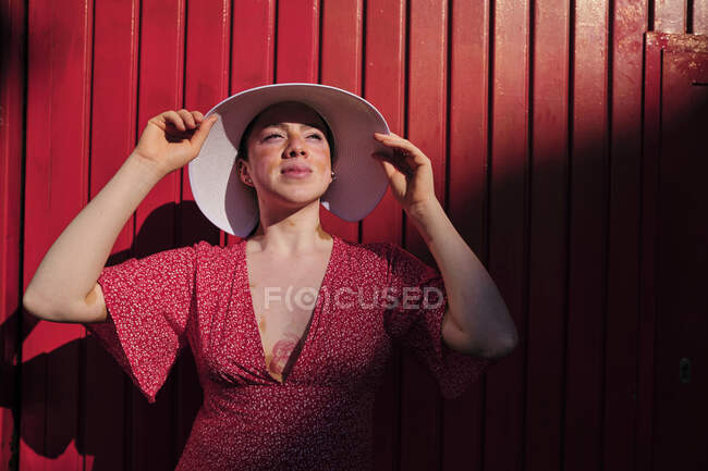 Mujer joven mirando hacia otro lado usando sombrero de sol por la pared roja - foto de stock