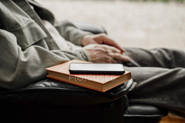 Mobile phone over book on chair by man at home — Stock Photo
