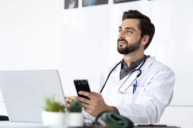 Médico masculino com telefone inteligente sentado na mesa enquanto olha para longe — Fotografia de Stock