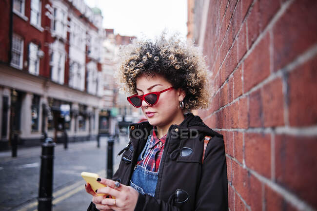 Jovem mulher usando óculos de sol usando telefone inteligente enquanto se inclina na parede durante as férias em Chinatown — Fotografia de Stock